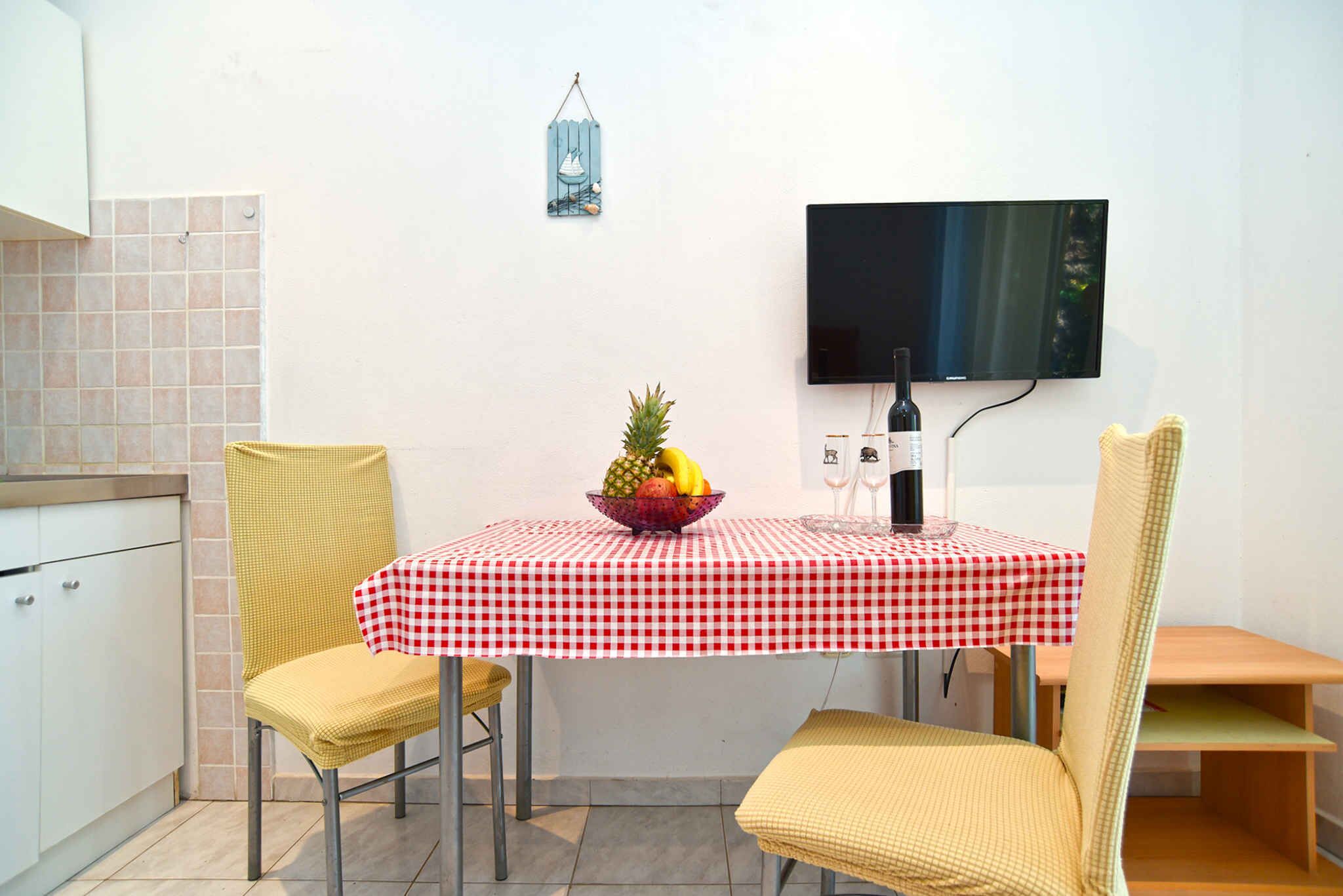 Un pequeño comedor con mantel a cuadros, dos sillas, un frutero, una botella de vino y un televisor montado en la pared.