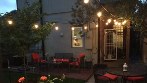 Un patio al aire libre con luces colgantes, asientos y un pequeño jardín con rosas.