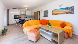 A modern living space with a kitchen, a large orange sofa, a wooden coffee table, and a small orange ottoman.