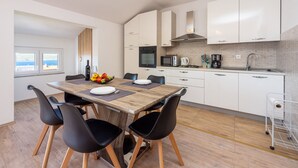 A modern kitchen with white cabinets, a wooden dining table, black chairs, and a view of the sea through a window.