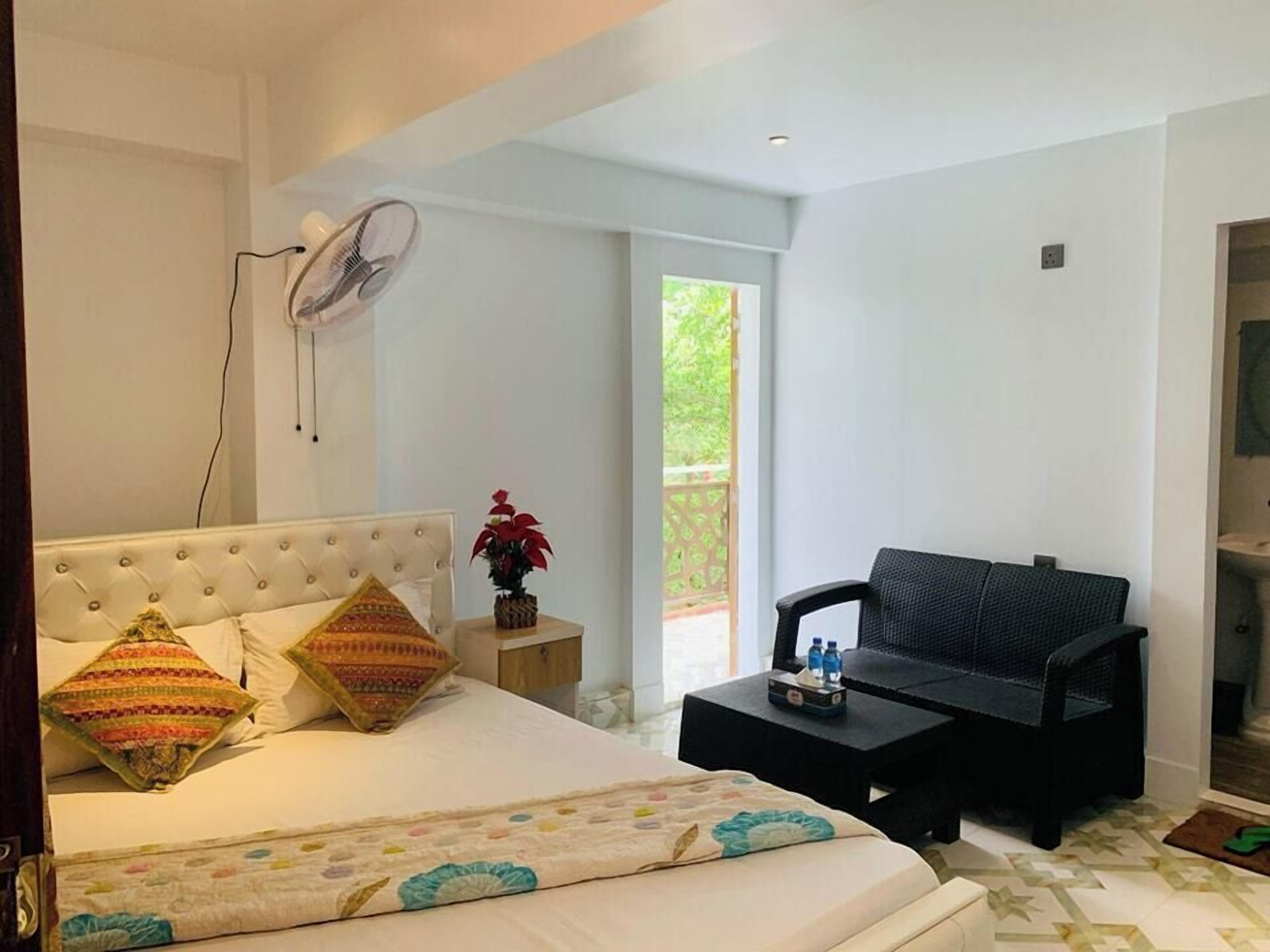 A bedroom with a bed, a bedside table with a fan, a window, and a view of greenery outside.