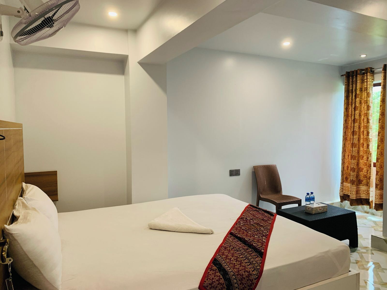 A hotel room with a bed, a chair, a small table with a bottle and tissues, and a fan.