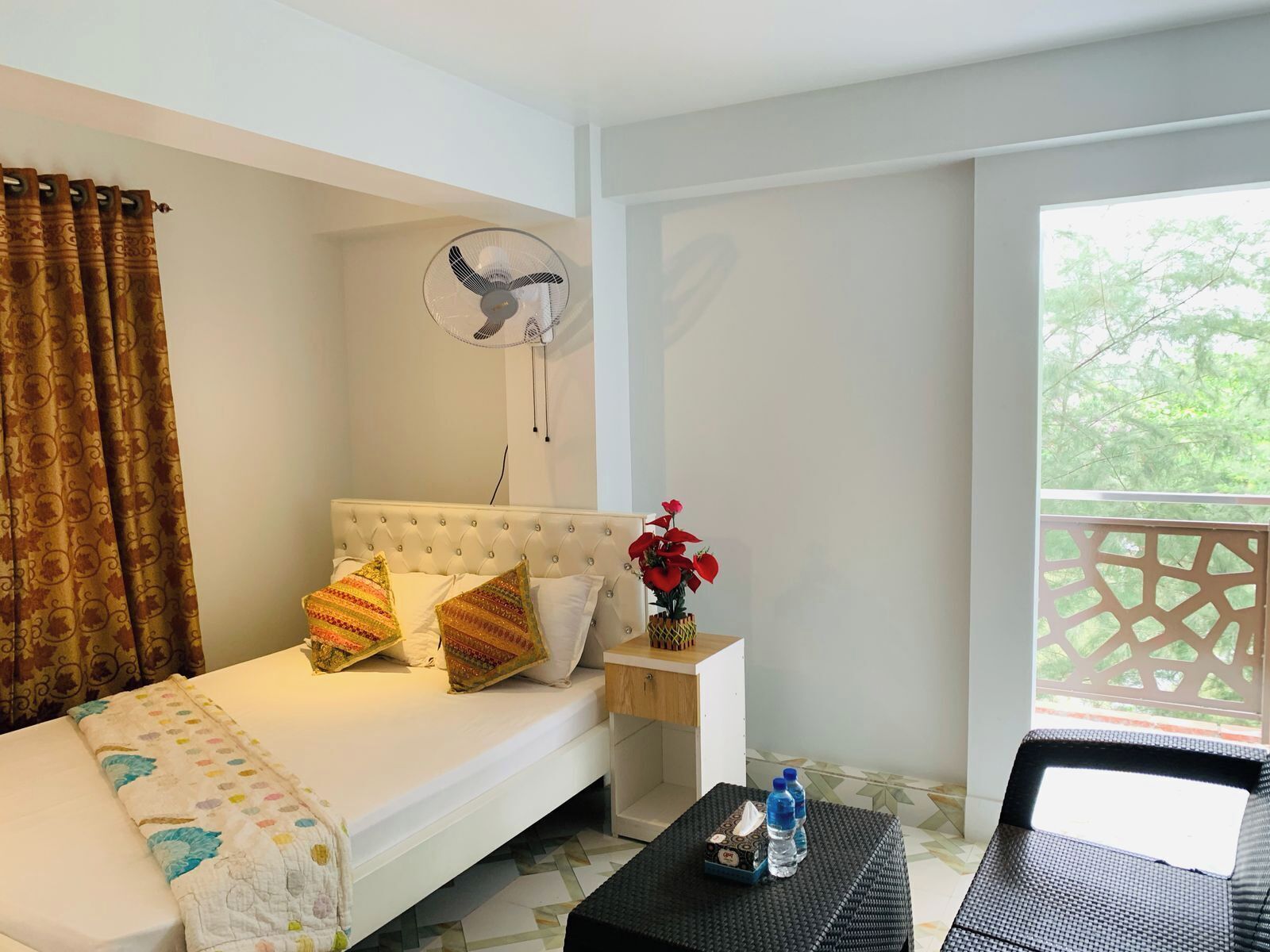 A room with a white sofa, patterned pillows, a bedside table with a vase of red flowers, a fan, and a view of greenery through a window.