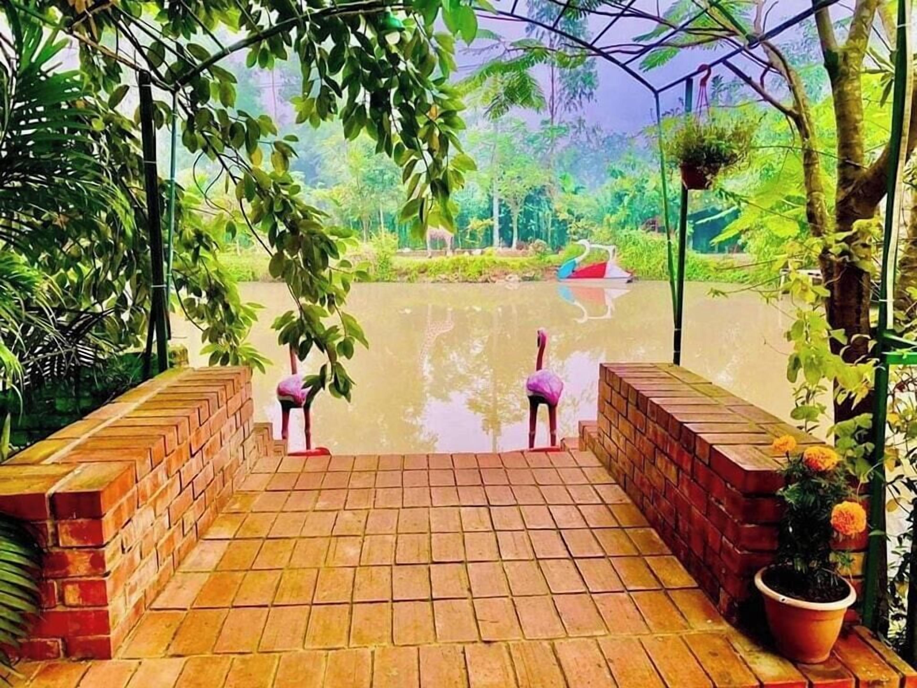 Ein Pflasterweg führt zu einem Gewässer, wo zwei Flamingos stehen, umgeben von Grünpflanzen und einem Blumentopf.