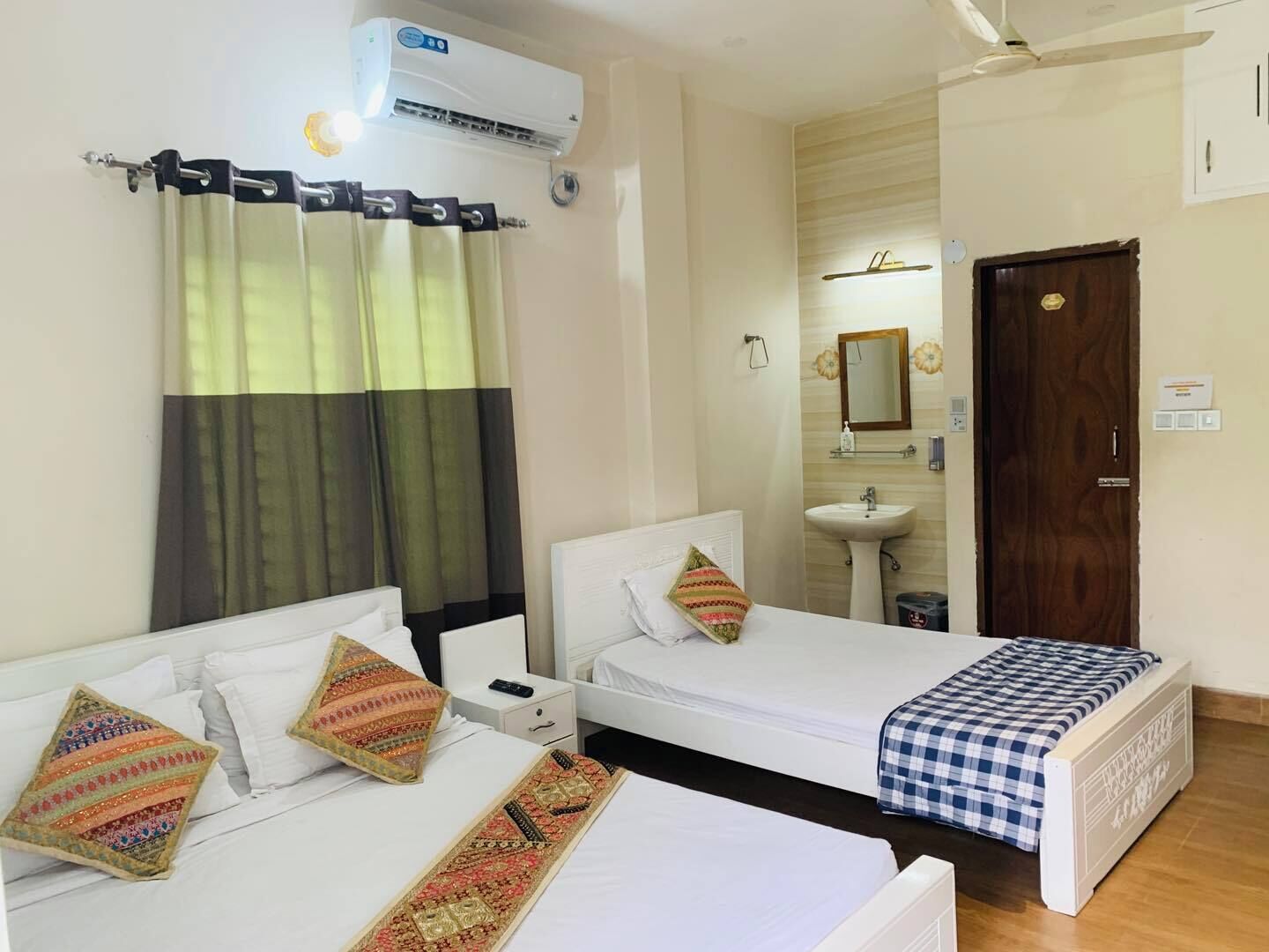 A hotel room with two single beds, a bathroom with a sink and mirror, and an air conditioning unit.
