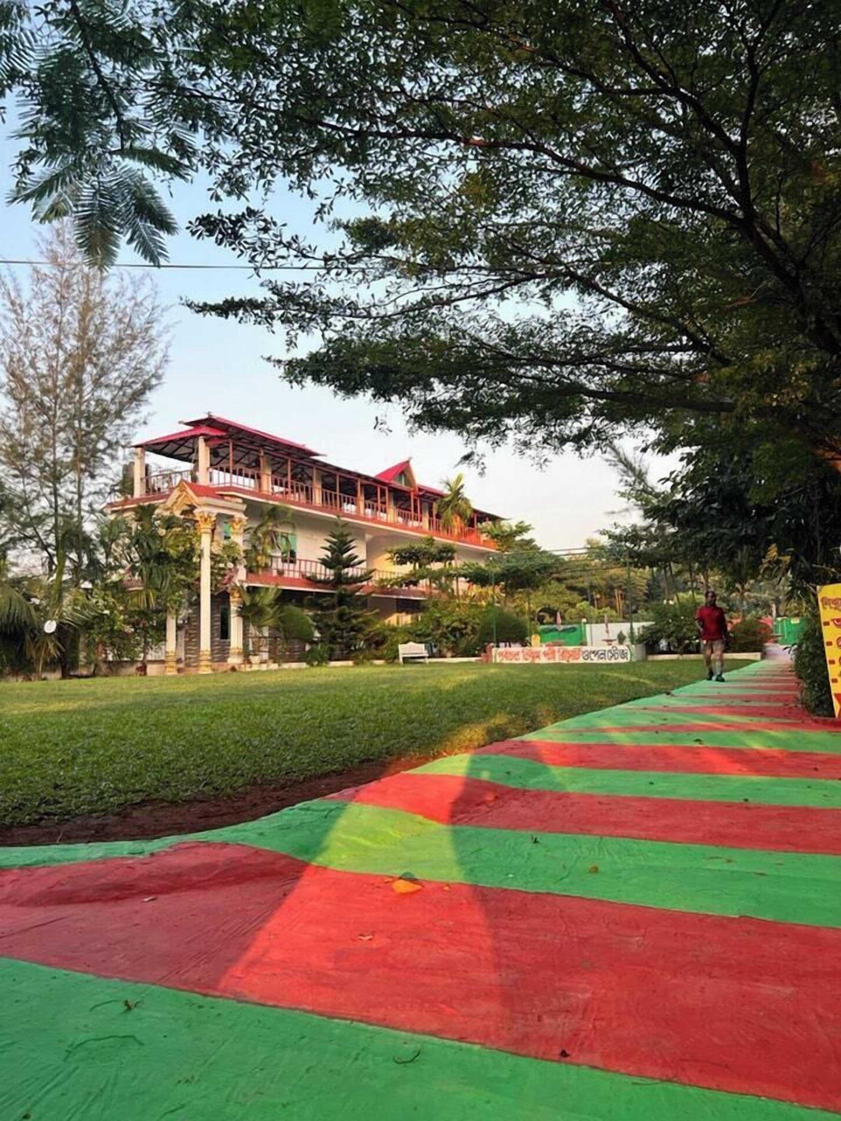 A building with a red roof and columns, surrounded by greenery and a pathway with red and green stripes.