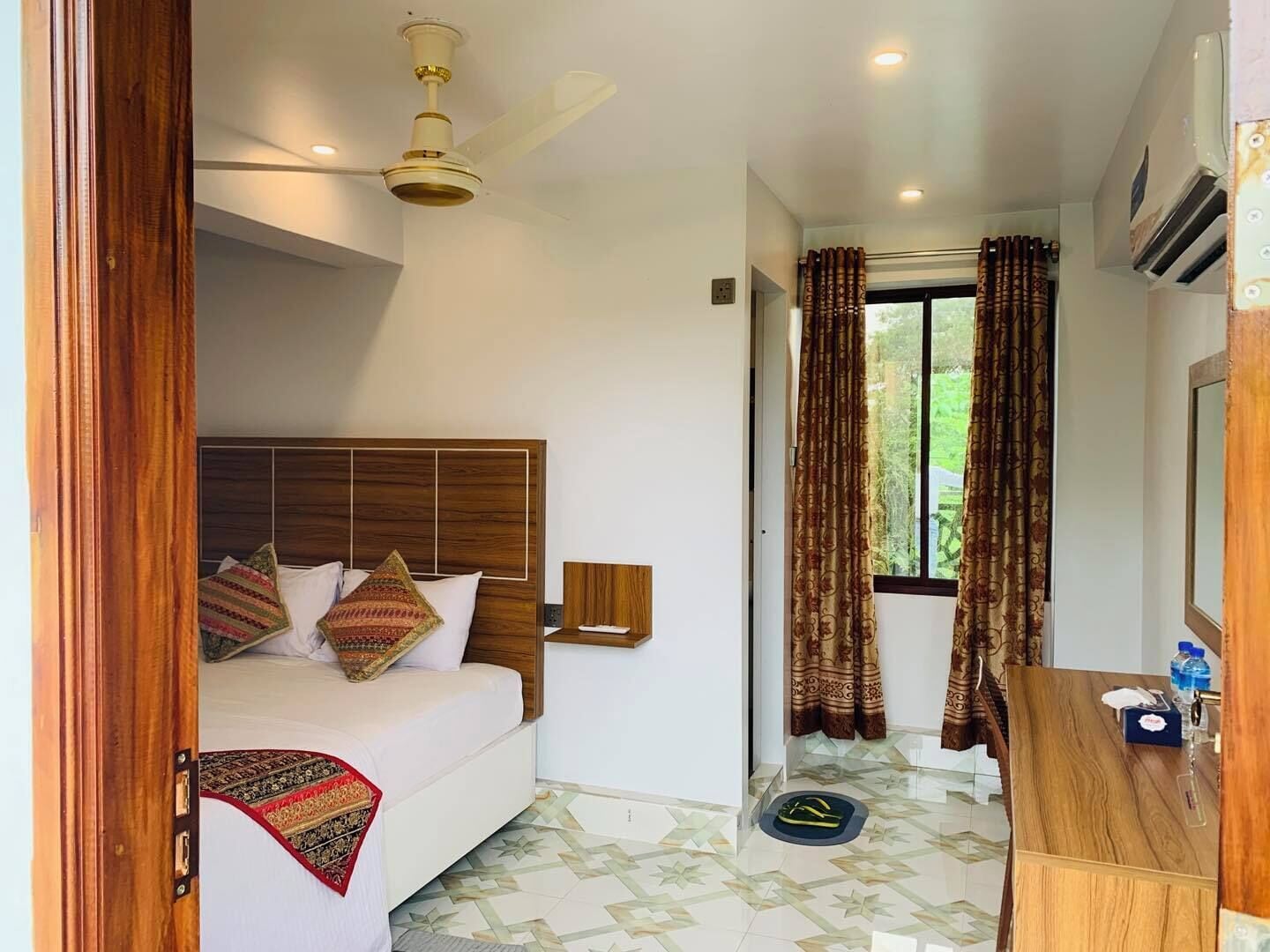 Una habitación de hotel con cama, escritorio, ventilador de techo y ventana con cortinas.