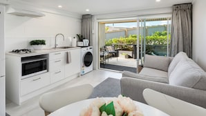 A modern kitchen with white cabinetry, a built-in oven, and a microwave. There is a dining area with a white table and chairs, and a grey sofa. A large window provides a view of the outdoors.