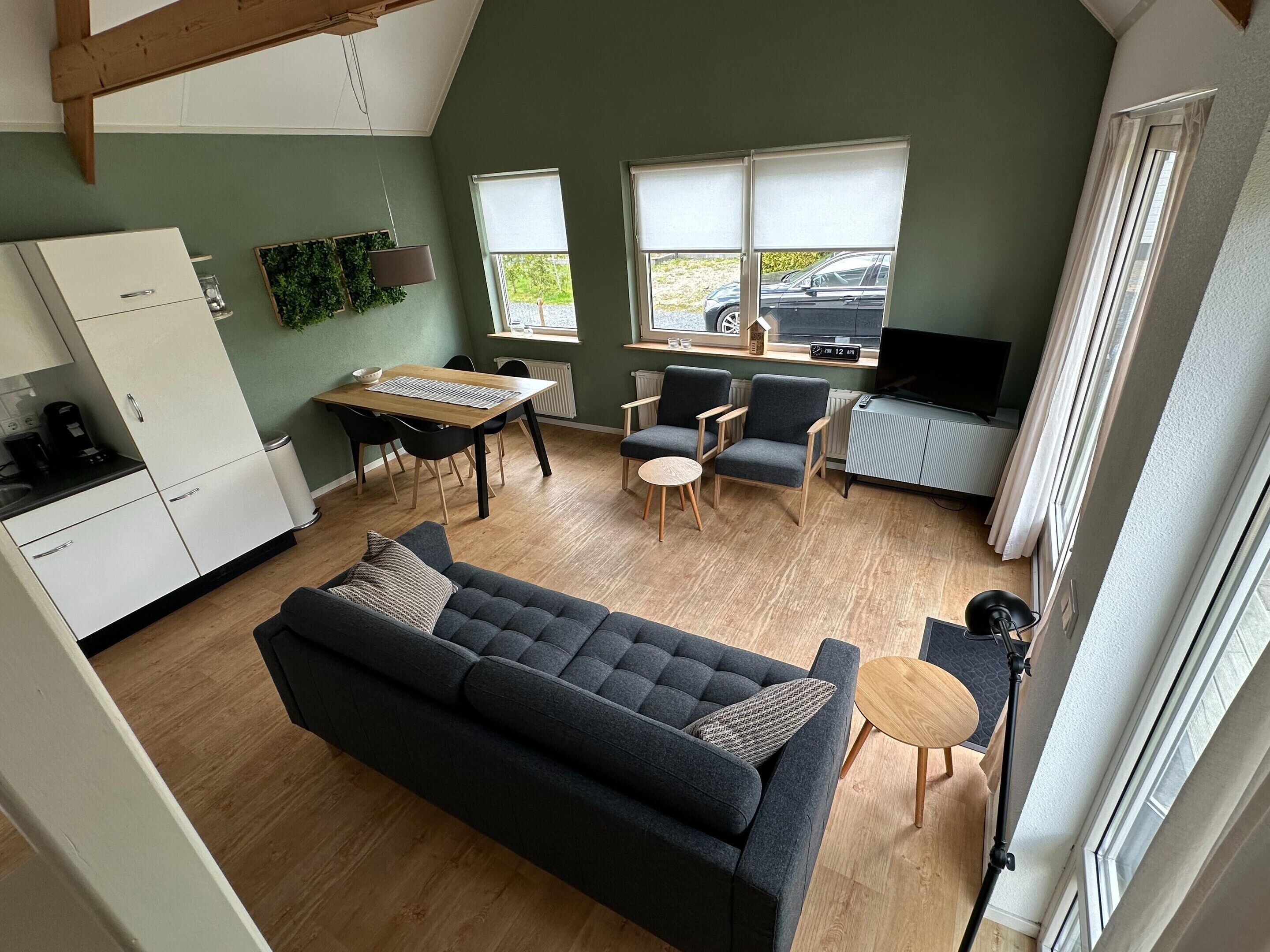A modern living room with a sofa, chairs, a dining table, a TV, and a kitchen area.