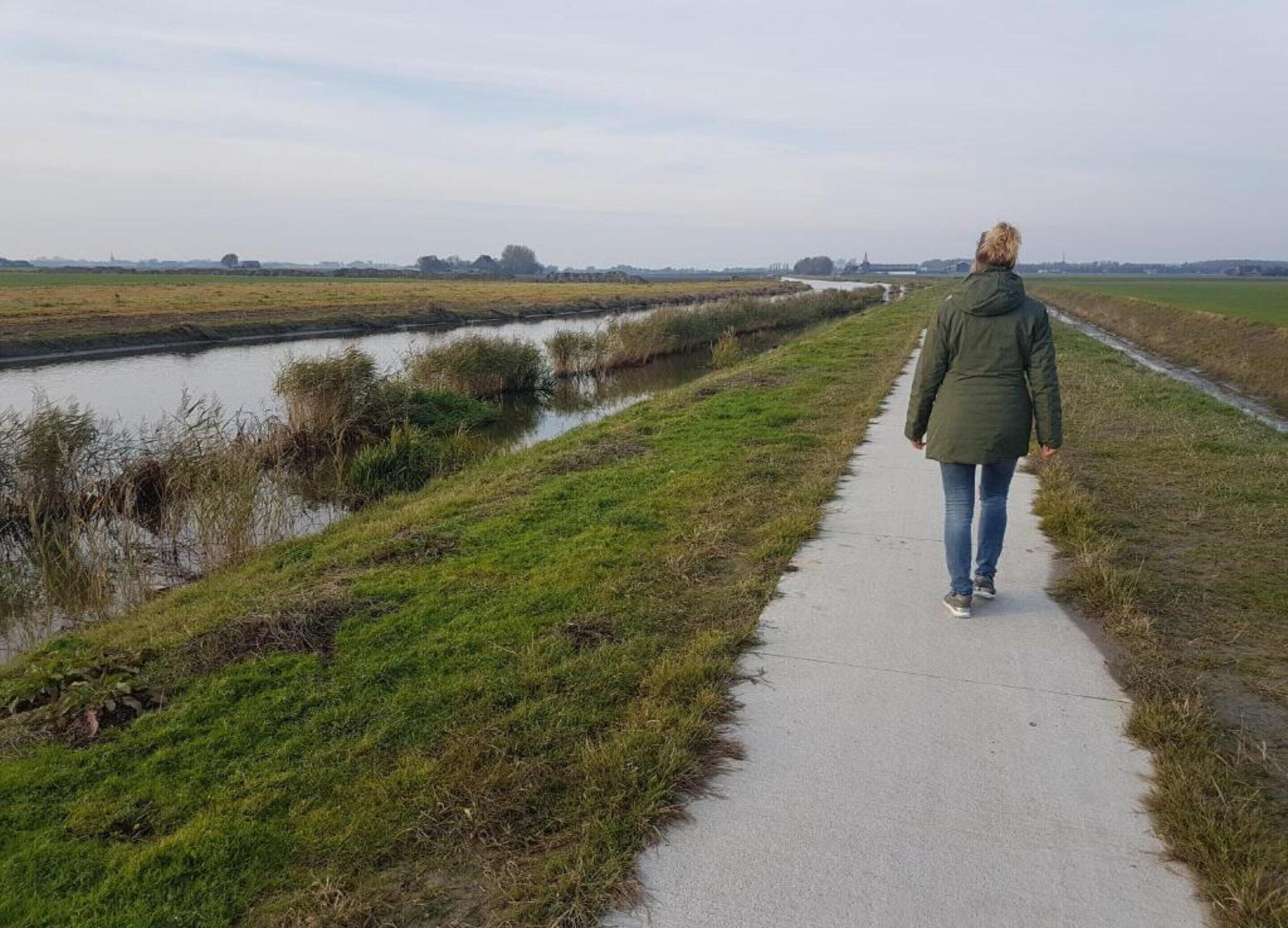 A person walking on a concrete path beside a canal with grassy banks and fields in the background.