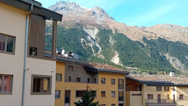 A multi-story building with balconies, a small tree in front, and a mountainous backdrop.