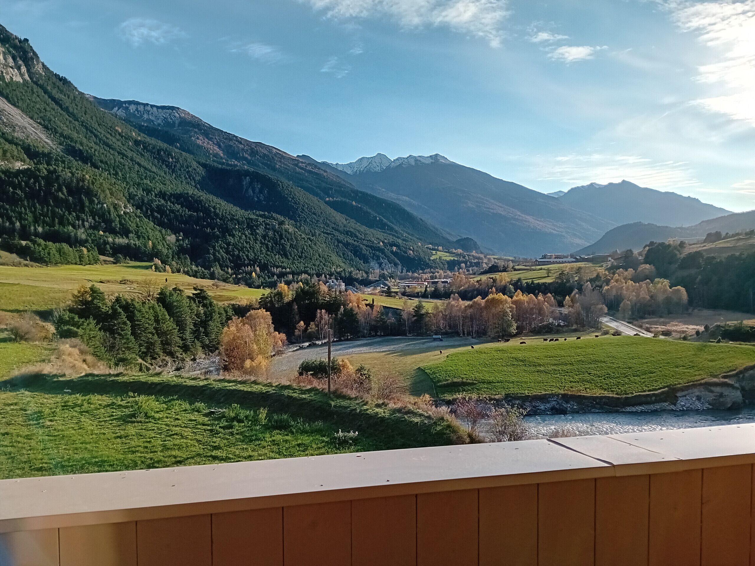 Eine gebirgige Landschaft mit einem Fluss, grünen Feldern und einem Gebäude in der Ferne.