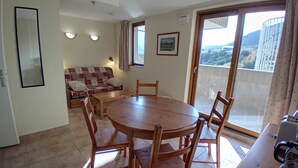 A dining area with a round wooden table and chairs, a sofa with cushions, and a view through a sliding glass door to an outdoor balcony.