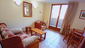 A living room with a sofa, a wooden table, and a chair. There is a window with curtains and a door leading outside.