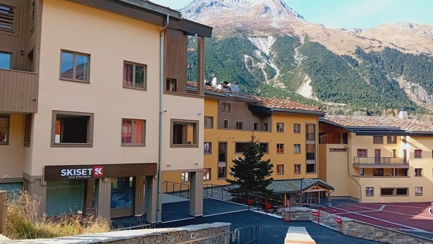 Un hotel de varios pisos con un cartel de estación de esquí, escalones de piedra y montañas al fondo.