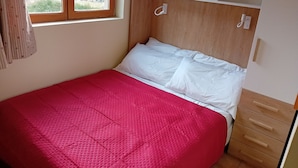 Un letto singolo con coperta rossa, testiera in legno e finestra vista alberi e campi.
