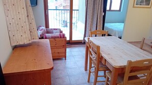 A room with a wooden bench, a dining table with chairs, a sofa with patterned cushions, and a view through a glass door to the outside.
