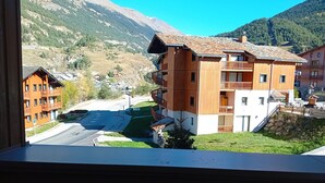 A multi-story building with balconies, a road in the foreground, and mountains in the background. - Terrasses E 305 - PARC NAT. VANOISE appart. 6 pers (Val Cenis)