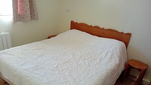 Een bed met een houten hoofdbord, een witte spreidek en een klein houten kruk ernaast.