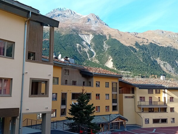A multi-story building with balconies, a small tree in front, and a mountainous backdrop.