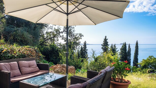 Ein Korbusofa mit Kissen, ein Tisch mit Glasplatte und ein großer Sonnenschirm in einem Patio-Bereich mit Blick auf das Meer und Bäume.