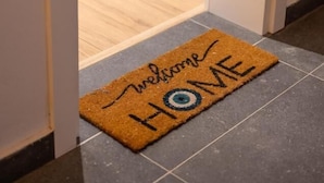 Eine braune Fußmatte mit der Aufschrift "Welcome Home" am Eingang eines Raumes mit Holzboden und gefliesten Wänden.