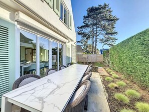 En uteplats med ett bord i marmor och flätade stolar, ett stort träd och en välklippt häck.