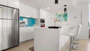A modern kitchen with white cabinets, a stainless steel refrigerator, a coffee machine, and a microwave. There is a small island with bar stools and a painting of a beach scene on the wall. - Aquaview Unit 604, 29 Canberra Tce, Kings Beach (Kings Beach)