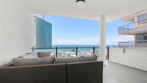A balcony with a wicker sofa, a view of the sea, and a modern building in the background. - Aquaview Unit 604, 29 Canberra Tce, Kings Beach (Kings Beach)