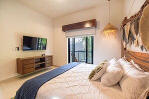 A modern bedroom with a large bed, a TV mounted on the wall, a wooden headboard, and a view of the outdoors through a sliding door. - Suite Tropical N11 Downtown (Cozumel)