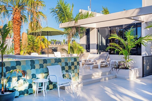 A modern outdoor lounge area with white lounge chairs, a green umbrella, and a stone wall with a mosaic design. - Suite Tropical N11 Downtown (Cozumel)