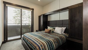 A bedroom with a large bed, a dark headboard, a sliding glass door, and a view of trees outside.