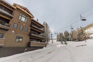 A multi-story building with balconies, a ski lift in the background, and a snowy slope.