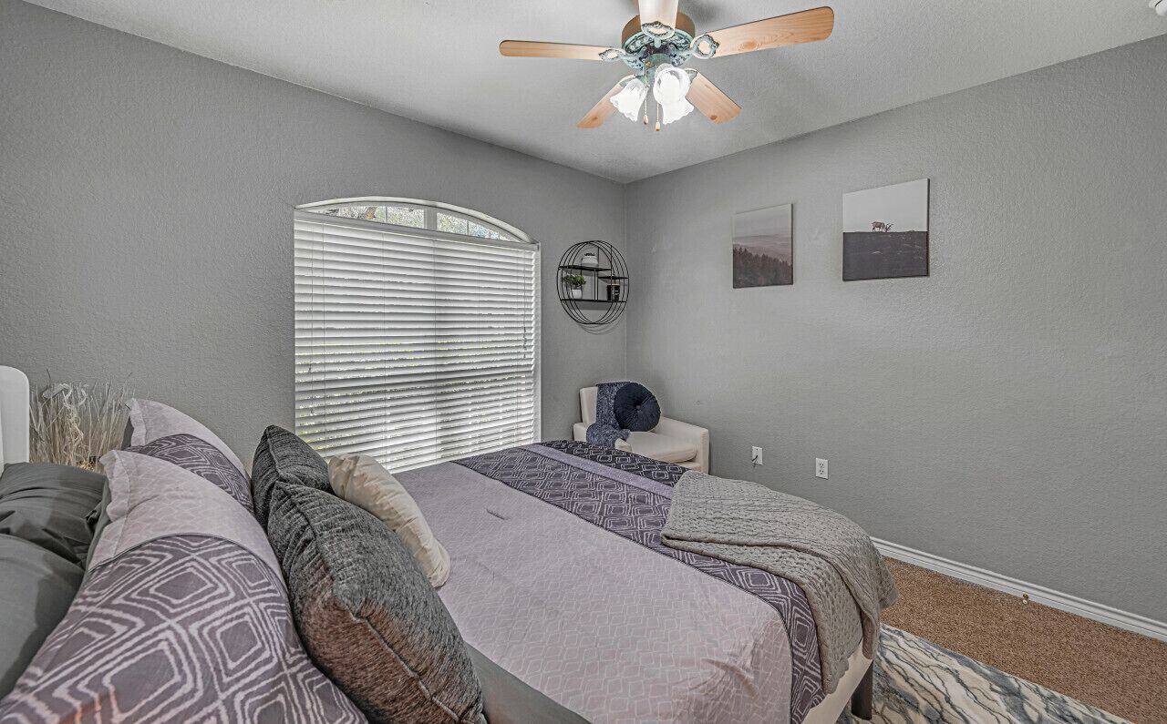 Une chambre à coucher comprenant un lit, un ventilateur de plafond, une fenêtre avec des stores et une table de chevet avec une lampe.