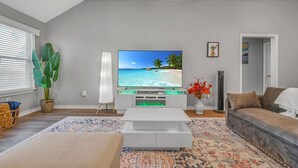 Ein Wohnzimmer mit Flachbildfernseher, weißem Couchtisch, gemustertem Teppich, Sofa und Topfpflanze.