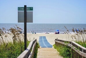 Una playa con un paseo marítimo de madera que lleva al océano, una señal de calle que dice 'CONCORD ST', y gente en la playa a lo lejos.