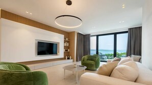 A modern living room with a flat-screen TV, green and beige sofas, a coffee table, and a large window offering a view of the sea. - Luxury Villa Beyond Waves with Pool (Baška Voda)
