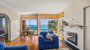 A living room with a blue sofa, wooden dining table, and chairs. There is a fireplace and a view of the ocean through large windows. - Oceans Edge - Spectacular home with ocean views on the hill of Mollymook Beach (Mollymook Beach)