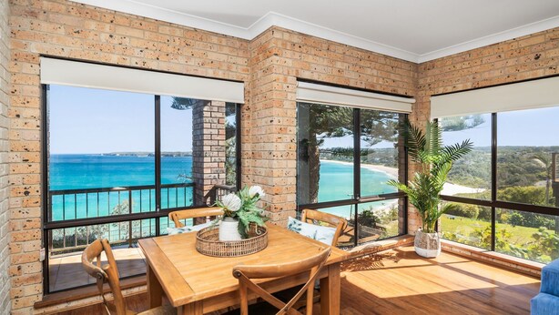 A room with a wooden table and chairs, large windows with a view of the sea, and a brick wall. - Oceans Edge - Spectacular home with ocean views on the hill of Mollymook Beach (Mollymook Beach)