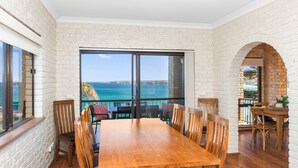 A dining area with a wooden table and chairs, large windows with a view of the sea, and a brick wall. - Oceans Edge - Spectacular home with ocean views on the hill of Mollymook Beach (Mollymook Beach)