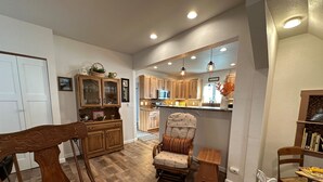 Un vaste salon doté d’un plancher en bois, un coin repas avec une table et des chaises en bois, une cuisine avec des meubles en bois, et un coin salon confortable avec un fauteuil rembourré et un petit tabouret en bois.