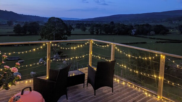 A wooden deck with wicker chairs and a table, overlooking a rural landscape with fields and trees. - The Barn @ Tyn y Ffridd , great views,
 WiFi, log burner (Denbighshire)