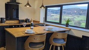 A modern kitchen with a wooden island, two bar stools, a large window with a view of the countryside, and a black range hood. - The Barn @ Tyn y Ffridd , great views,
 WiFi, log burner (Denbighshire)