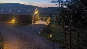 A rural property with a paved driveway leading to a gated entrance, a small illuminated structure, and a view of a hilly landscape in the background. - The Barn @ Tyn y Ffridd , great views,
 WiFi, log burner (Denbighshire)