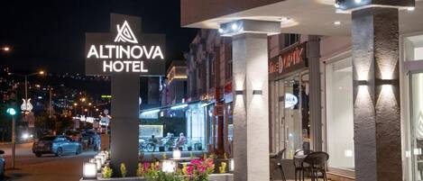 The exterior of the Altinova Hotel with a prominent sign, outdoor seating area, and a view of the street at night.
