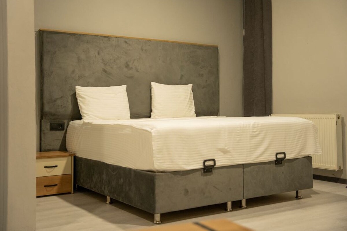 A bedroom with a grey bed, white pillows, a wooden nightstand, and a radiator.