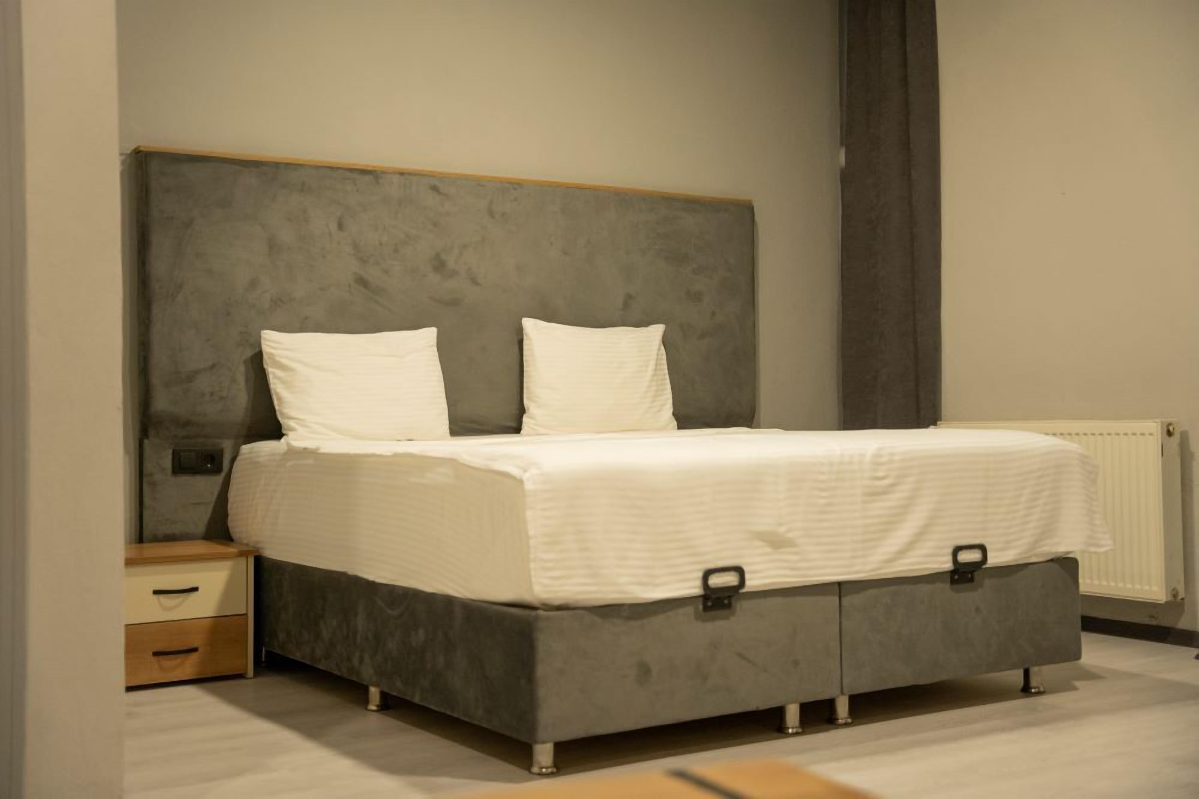 Une chambre à coucher avec un lit recouvert d’un tissu gris, deux oreillers blancs, une table de chevet en bois avec des tiroirs et une tête de lit grise.