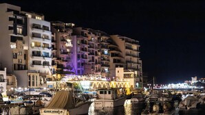 Un port de plaisance avec des bateaux amarrés, un bâtiment aux couleurs vives doté de balcons, et une vue nocturne sur la ville en arrière-plan.