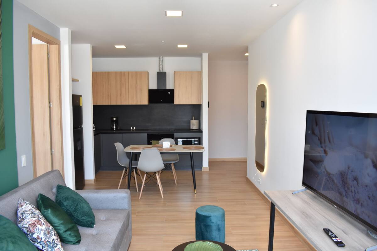 Uma sala de estar moderna com um sofá cinzento, almofadas verdes, uma televisão e um espaço de refeições com mesa e cadeiras.