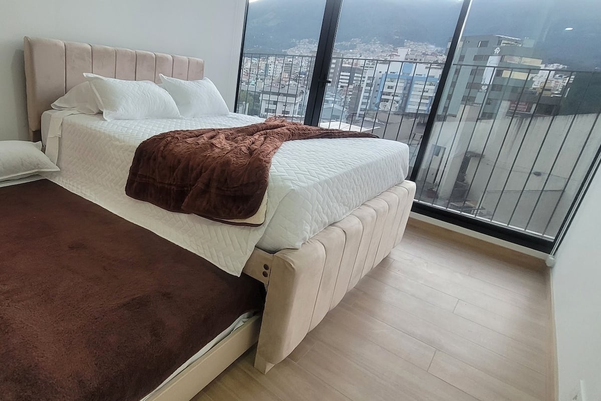 Quarto com uma cama grande, um cobertor marrom e vista para a cidade através de uma varanda.